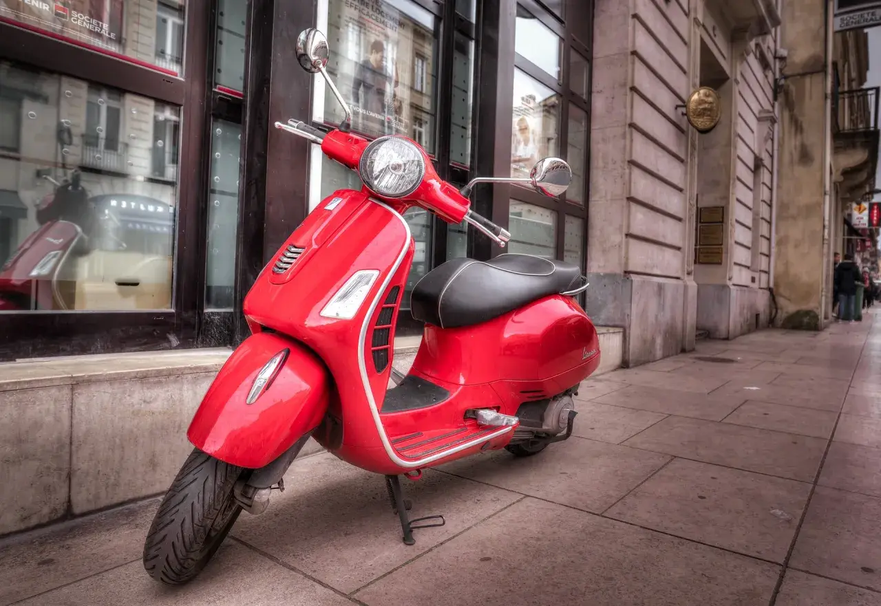 Czerwony skuter Vespa, idealny do zdobycia prawa jazdy AM, stoi na chodniku przed budynkiem.