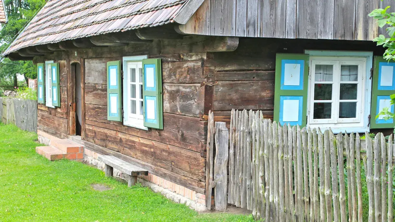 Drewniana chata z zielono-niebieskimi okiennicami, ławką i płotem. Skansen w Dziekanowicach zaprasza do podróży w czasie.