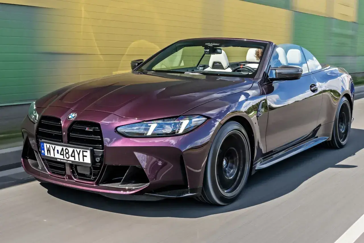 Fioletowe BMW M4 Competition kabriolet pędzi po drodze, odsłaniając białe wnętrze i sportowe detale.