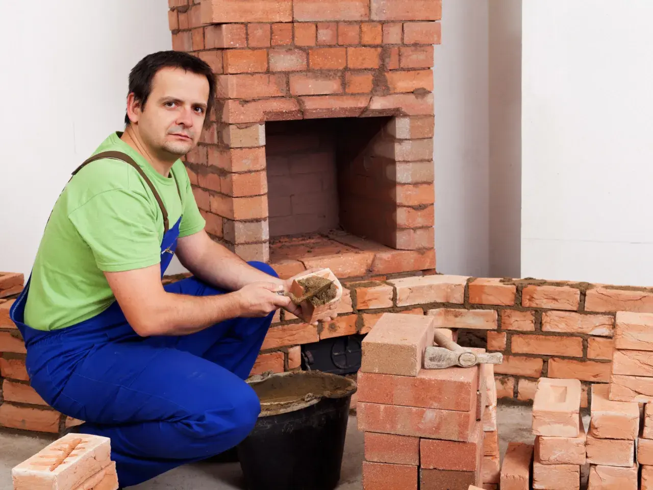 Murarz układa cegły szamotowe, budując kominek. W wiadrze zaprawa, obok stosy cegieł.