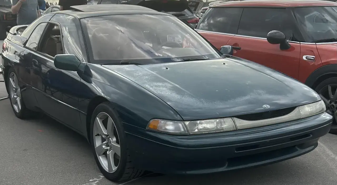 Subaru SVX w ciemnozielonym kolorze, z charakterystycznymi oknami w stylu "glass-canopy", zaparkowany obok czerwonego samochodu.