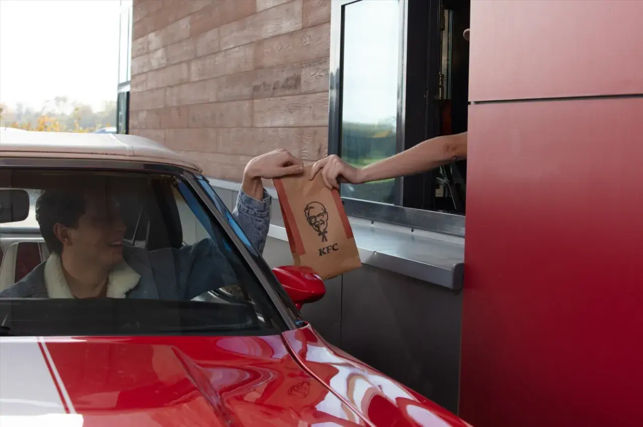 Mężczyzna w czerwonym aucie odbiera z okienka KFC torbę z jedzeniem. Wygląda na zadowolonego, że otrzymał swój mega pocket kcal.