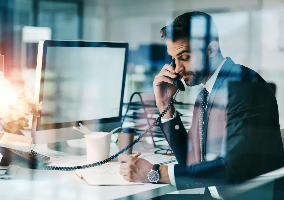 Biznesmen w garniturze rozmawia przez telefon, notując w zeszycie. Nawet w obliczu siły wyższej, praca wre.