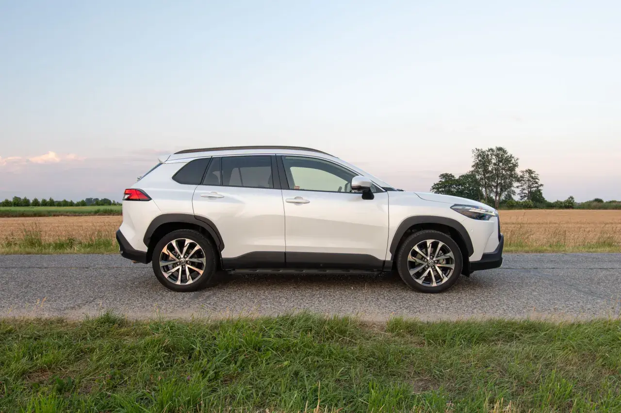 Biała Toyota Corolla Cross Hybrid stoi na poboczu drogi, otoczona polami i zieloną trawą.