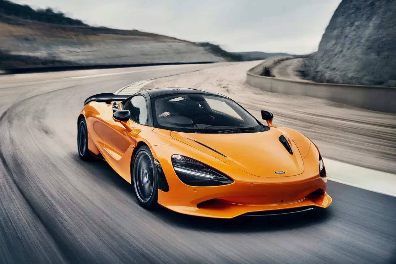 Pomarańczowy McLaren 750S z czarnym dachem sunie po torze wyścigowym, tworząc dynamiczny obraz prędkości i mocy.