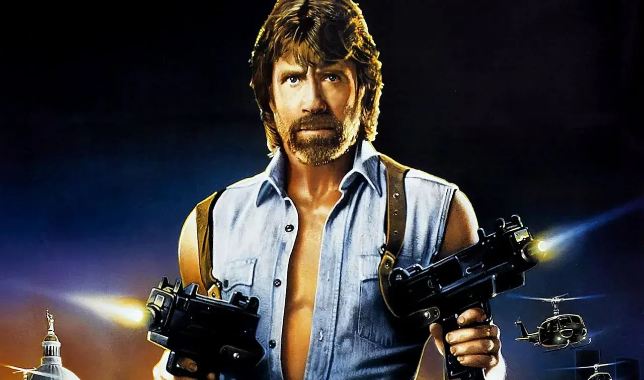 Chuck Norris z karabinami w akcji, jak w jego najlepszych filmach. W tle helikoptery i budynek.