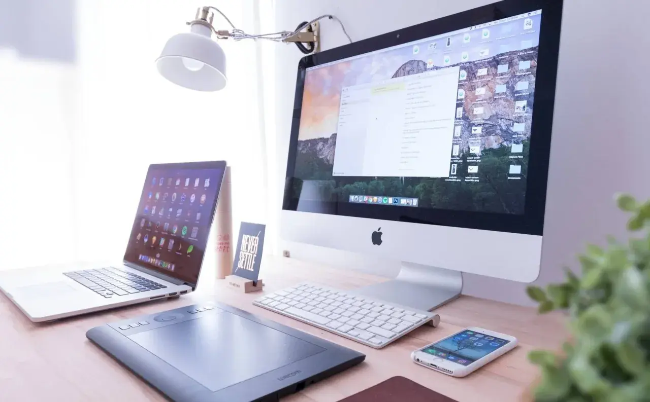 Biurko z iMac, laptopem, tabletem graficznym i telefonem. Na ekranie komputera widać okno programu Adobe Dreamweaver, gotowe do pracy nad stroną.