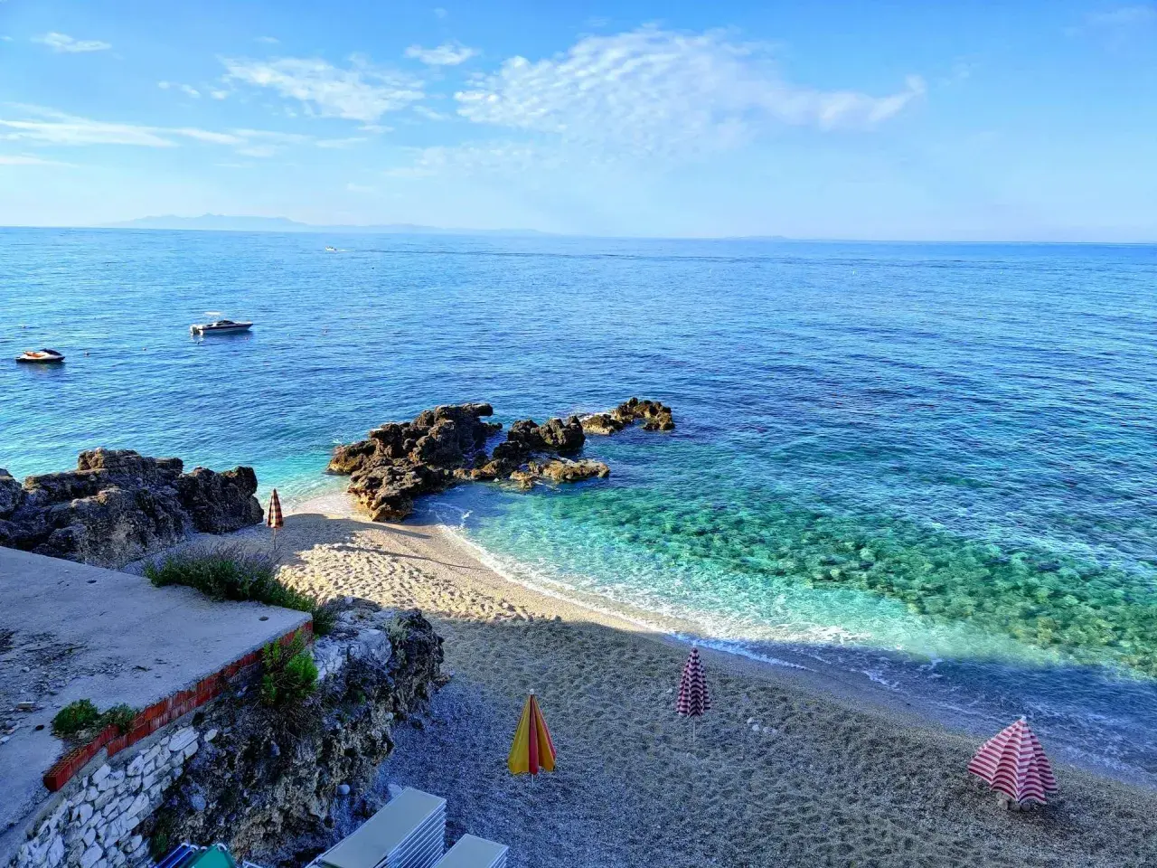 Albania, czerwiec: słoneczna pogoda nad krystalicznie czystym morzem, kamienista plaża z parasolami i łódkami na horyzoncie.