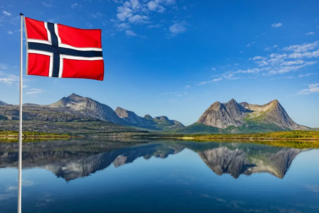 Norweska flaga powiewa nad jeziorem, odbijając się w wodzie. Czy do Norwegii potrzebny jest paszport? Piękne góry w tle.