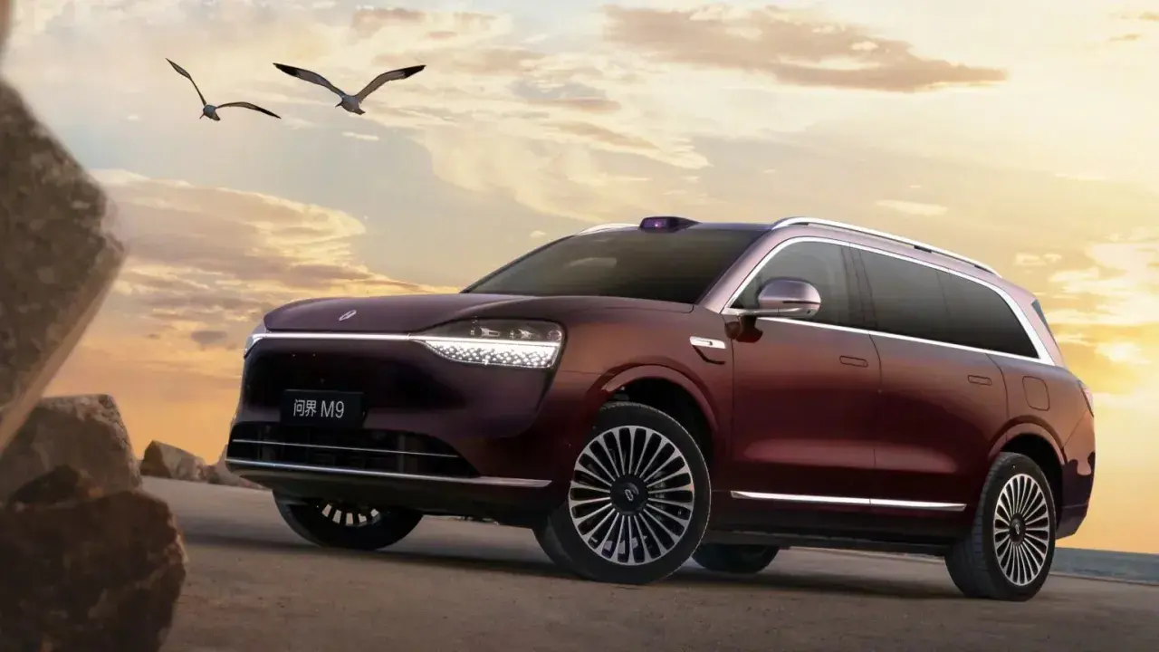 Ciemnoczerwony SUV Aito M9 na tle zachodzącego słońca, z ptakami w locie.