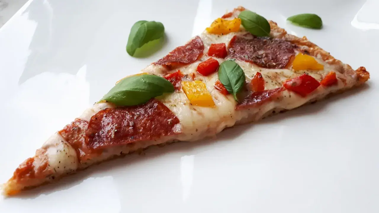 Kawałek pysznej pizzy keto z salami, papryką i bazylią na białym talerzu.