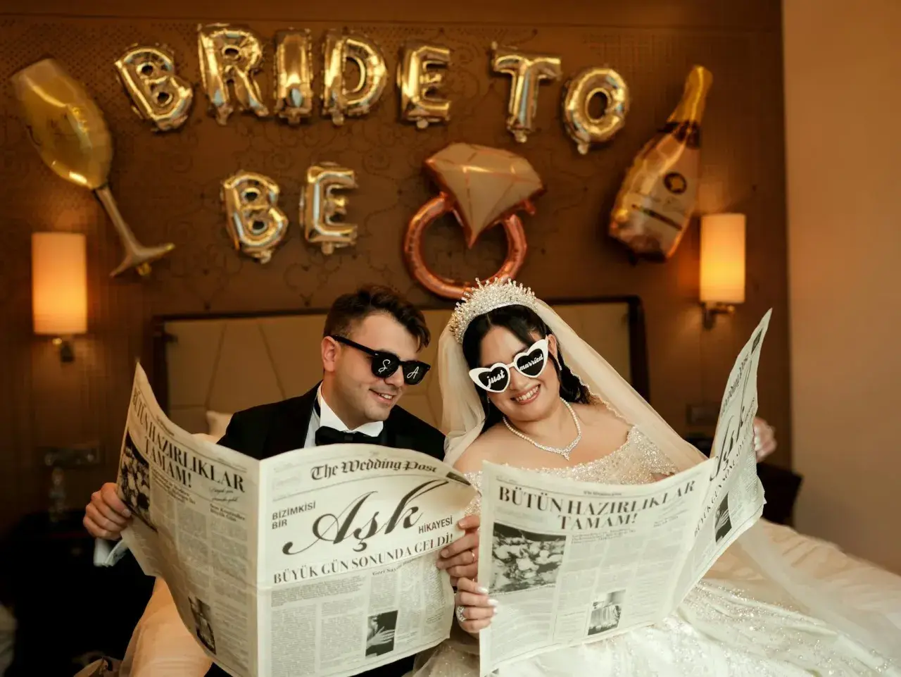 Para Młoda czyta gazetę z nagłówkiem "przykładowy harmonogram wesela". Balony "Bride to be" i kieliszek szampana w tle.