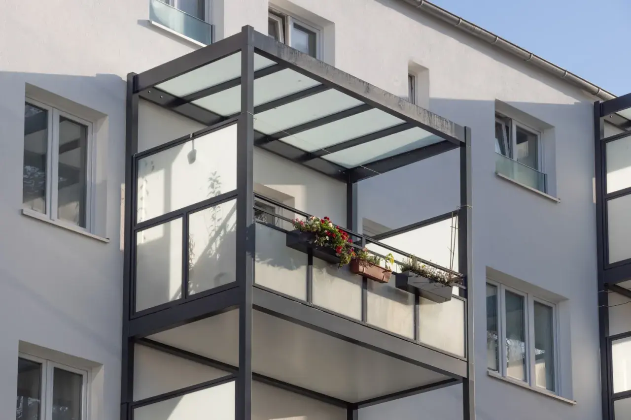 Nowoczesny balkon dostawny z przeszklonym zadaszeniem i balustradą, ozdobiony skrzynkami z kwiatami. Sprawdź balkon dostawny cena.