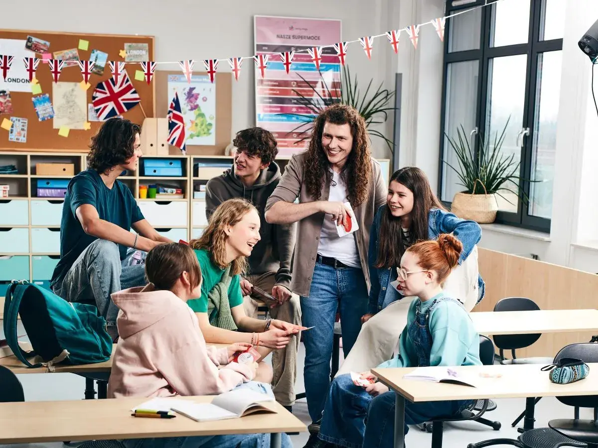 Uśmiechnięty nauczyciel z długimi włosami rozdaje karty uczniom w klasie. Wszyscy wyglądają na zaangażowanych w grę.