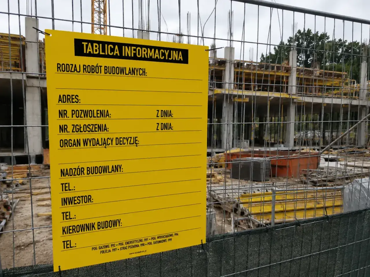 Żółta tablica informacyjna budowy z danymi o pozwoleniu, adresie i kontaktach. W tle widać konstrukcję budynku.