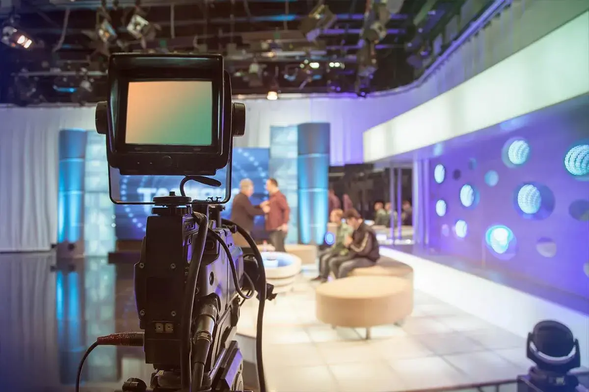 Telecamera in primo piano in uno studio televisivo. Si intravede il backstage, con persone sedute e in piedi, luci soffuse e un'atmosfera di attesa.