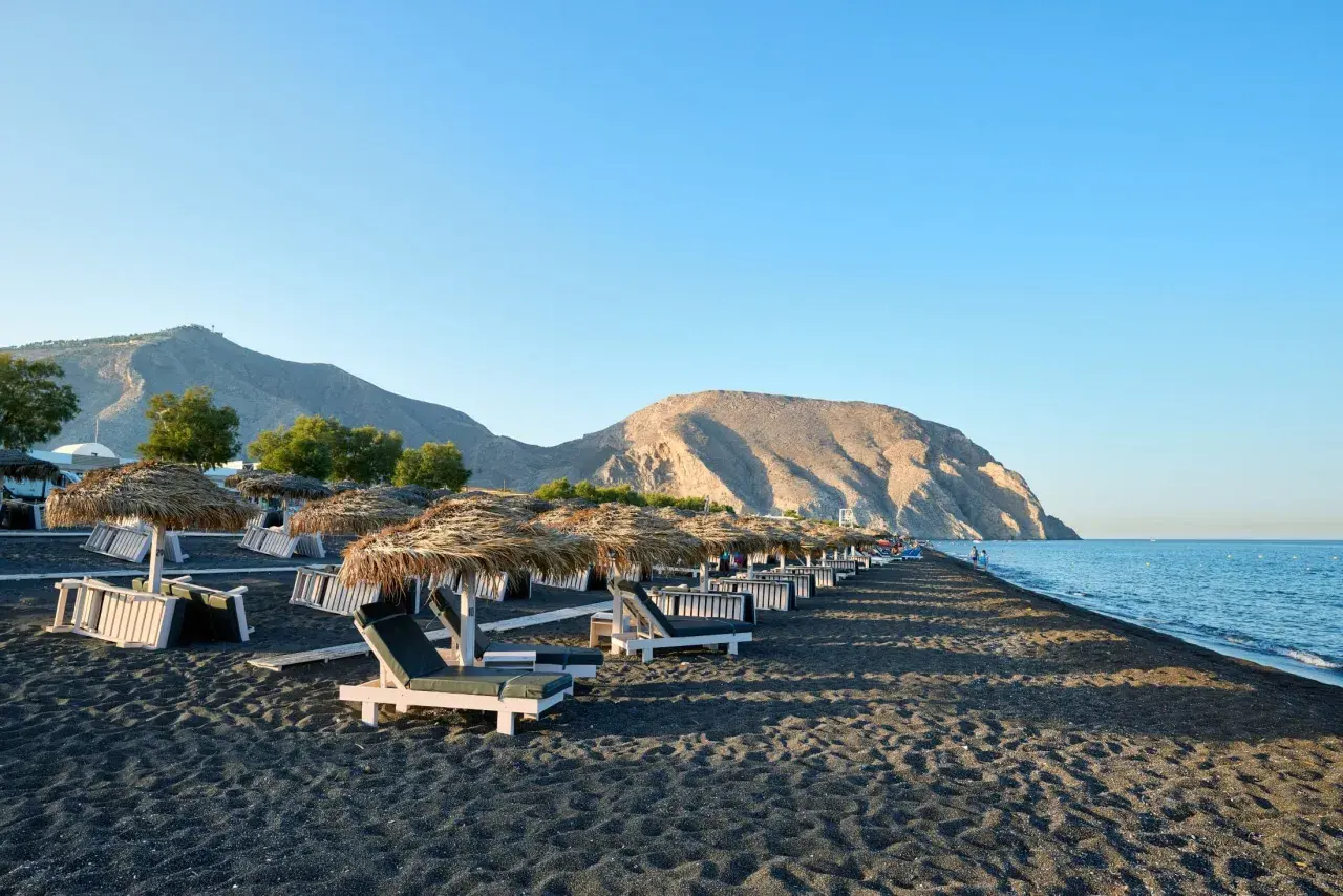 Czarne piaski i leżaki pod słomianymi parasolami na Santorini plażach. W tle góry i spokojne morze.