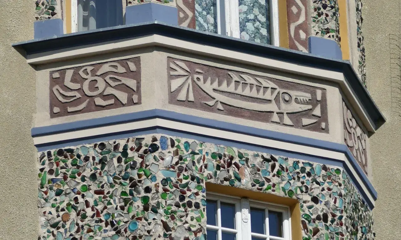 Sgraffito na fasadzie budynku: stylizowana ryba i abstrakcyjne wzory. Dolna część ozdobiona mozaiką z kolorowego szkła.
