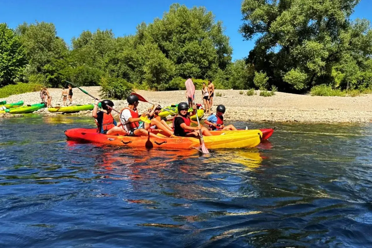 Des personnes profitent d'une activité canoe kayak sur une rivière ensoleillée, entourées de verdure et d'une plage de galets.