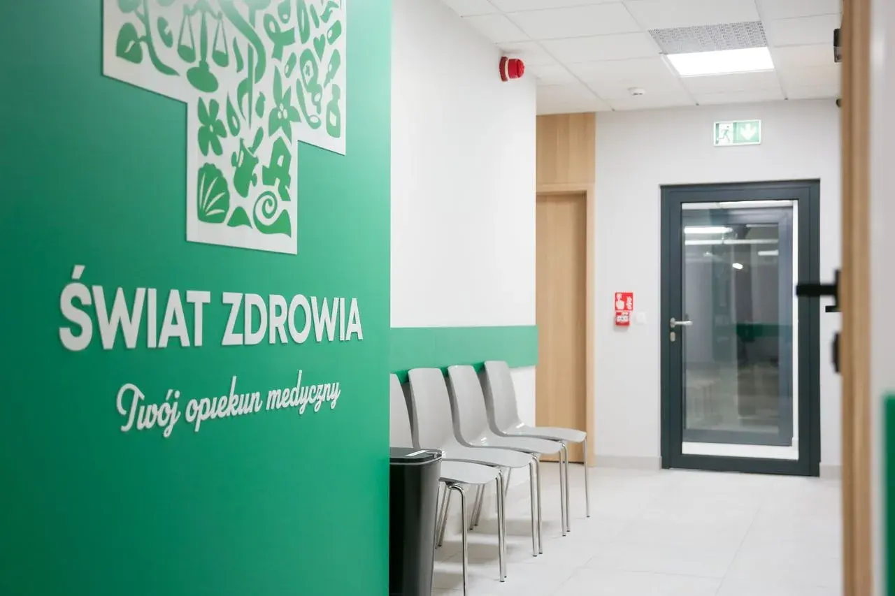 Zielona ściana z napisem "Świat Zdrowia", rząd krzeseł i drzwi w poczekalni kliniki.