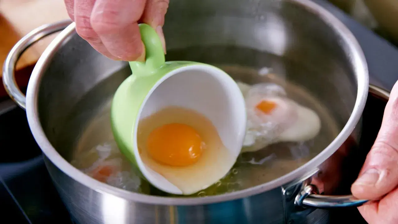 Ein rohes Ei wird aus einer grünen Tasse in kochendes Wasser gegossen, um verlorene Eier zuzubereiten.