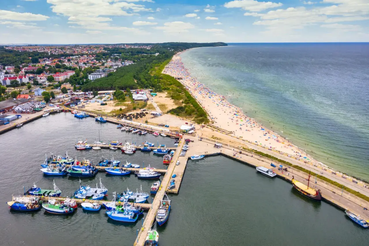 Port rybacki we Władysławowie, województwo pomorskie, tętni życiem. W tle rozciąga się plaża pełna turystów i morze.