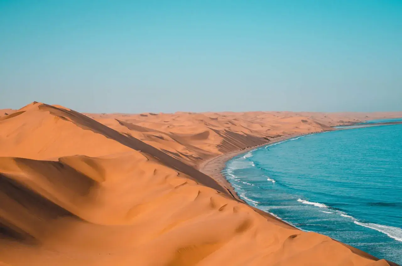 Piaskowe wydmy Namibii spotykają się z turkusowym oceanem. Idealna namibia pogoda na podziwianie tego kontrastu.