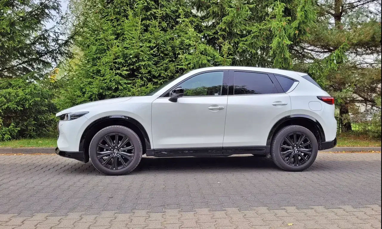 Nowa Mazda CX-5 w białym kolorze, zaparkowana na brukowanym podjeździe, otoczona zielenią drzew.