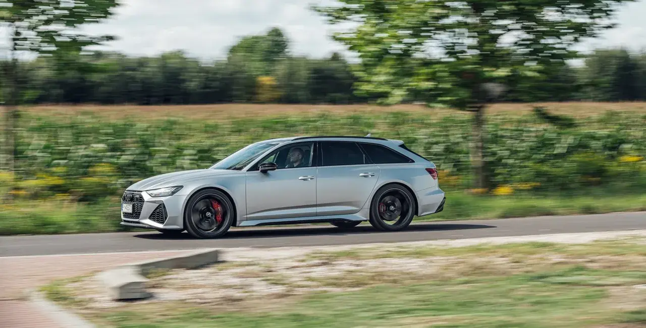 Srebrne Audi RS6 Avant pędzi po wiejskiej drodze, otoczone zielenią pól i drzew.