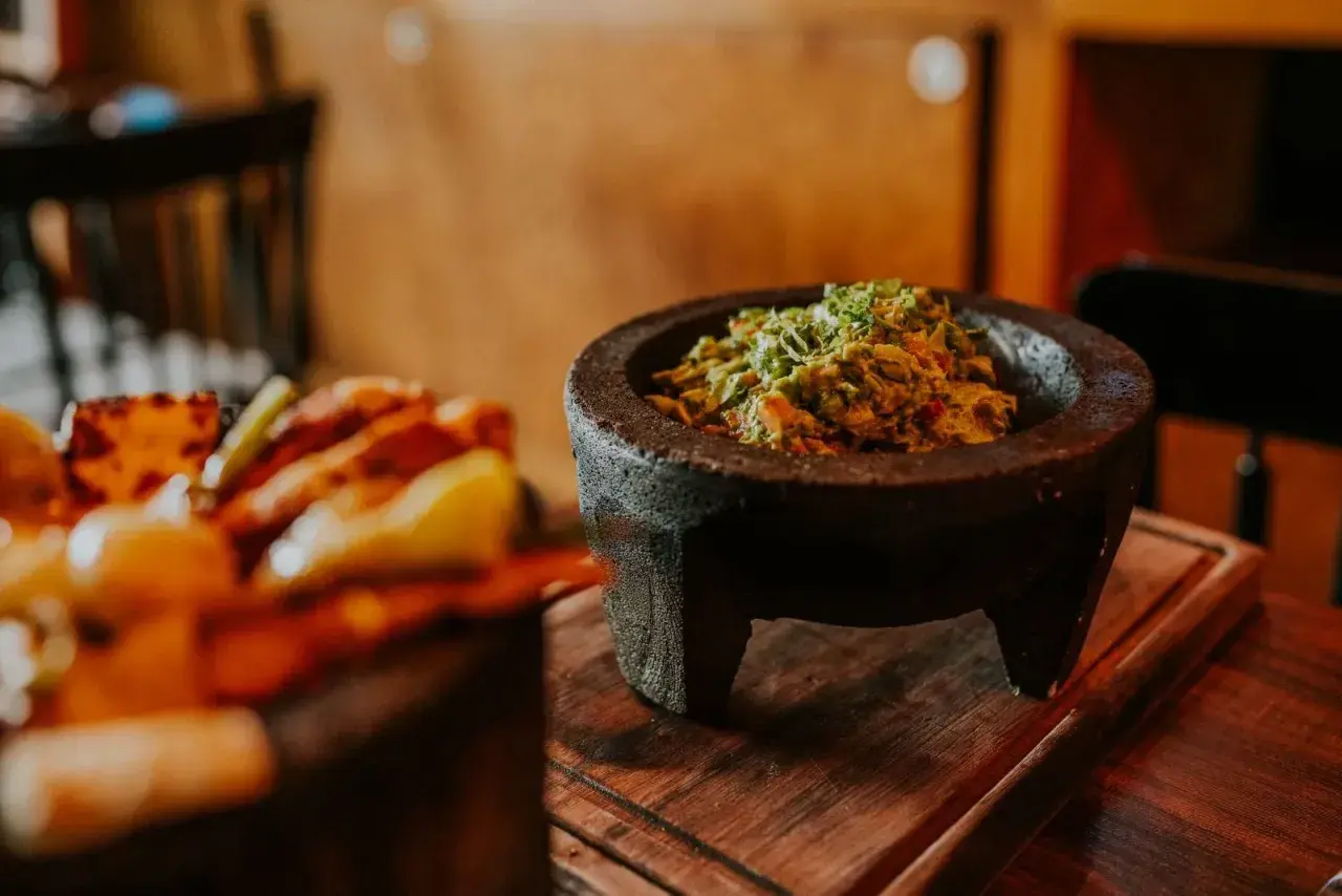 Świeże guacamole w kamiennym moździerzu, obok talerza z innymi najpopularniejszymi daniami meksykańskimi.