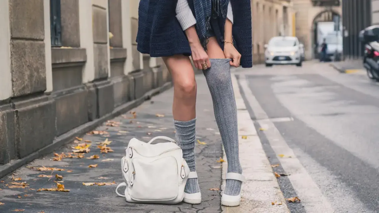 Stylizacja z długimi skarpetkami damskimi, biały plecak i jesienne liście na chodniku.