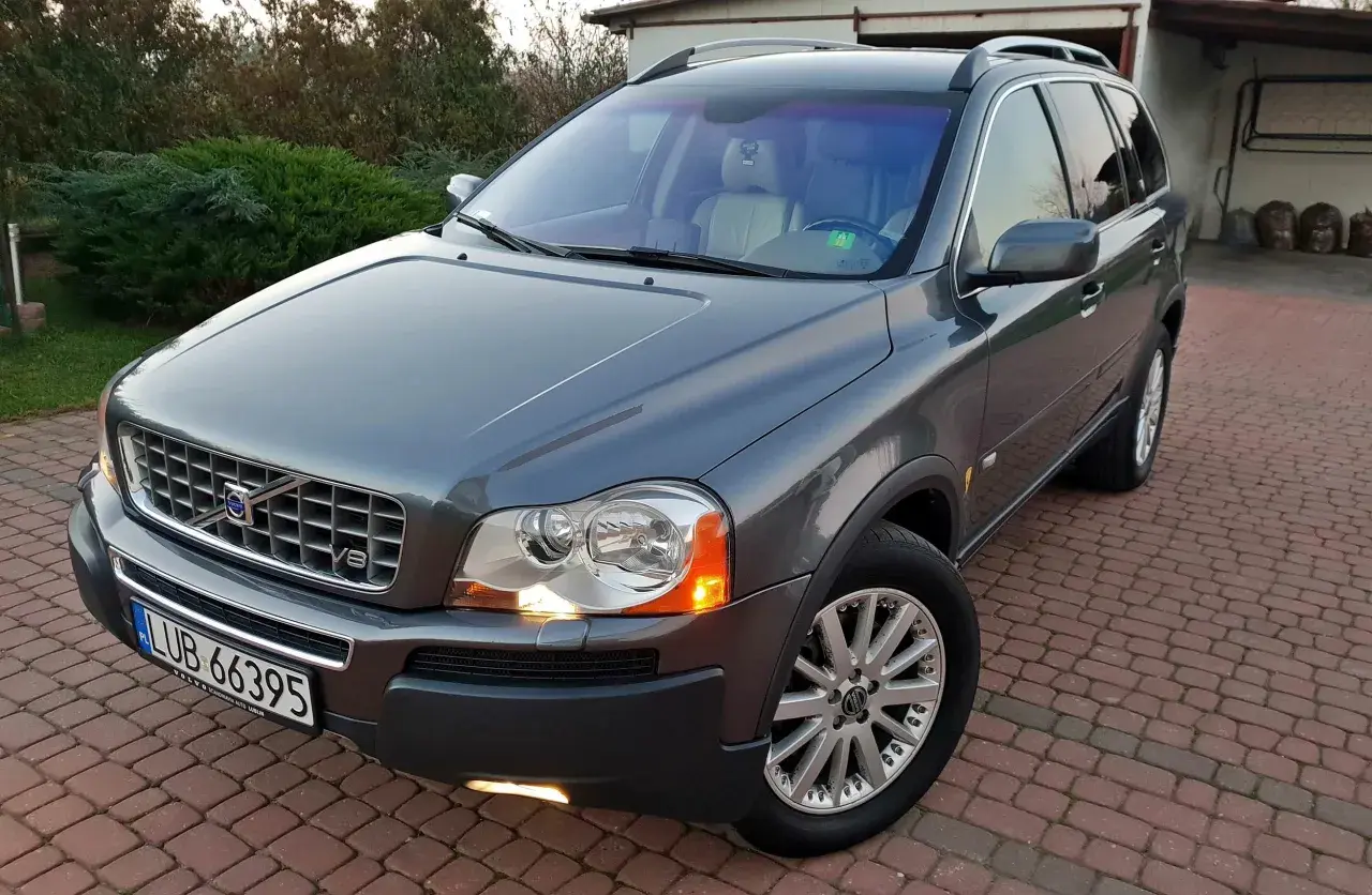 Szary Volvo XC90 z silnikiem V8, zaparkowany na podjeździe z kostki brukowej, otoczony zielenią.
