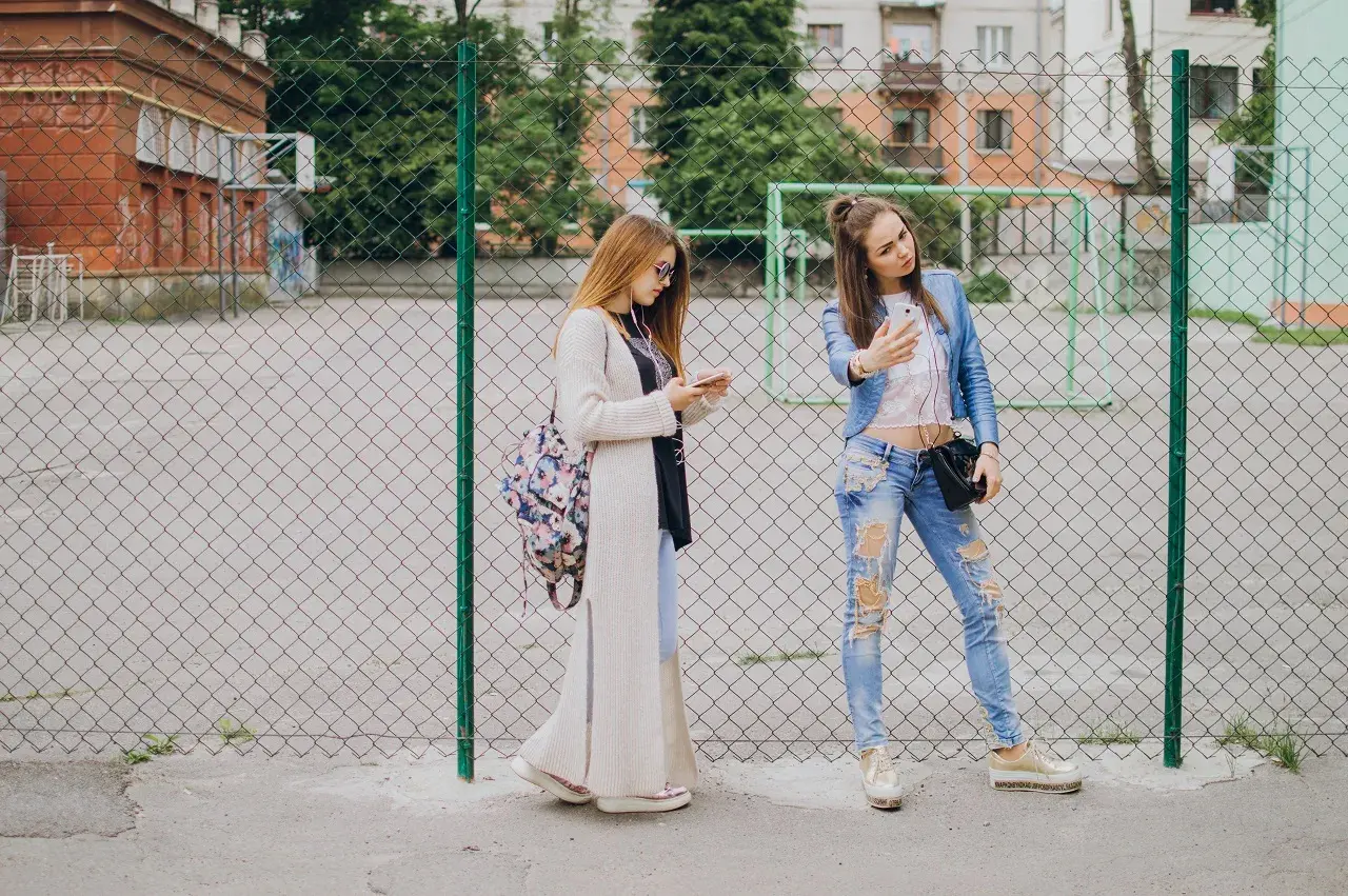 Dwie dziewczyny w modnych stylizacjach, idealnych na outfit do szkoły, stoją przy płocie. Jedna robi sobie selfie, druga przegląda telefon.