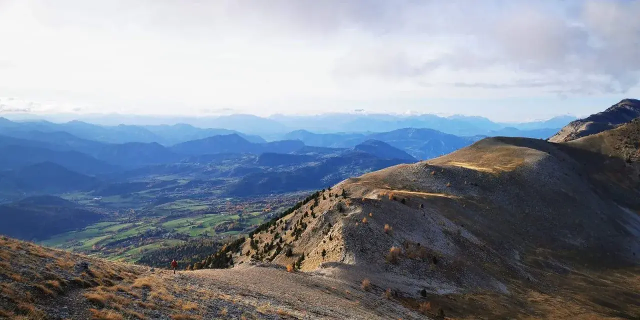 Un randonneur solitaire entame sa grande traversée des Alpes, dominant une vallée verdoyante sous un ciel nuageux.