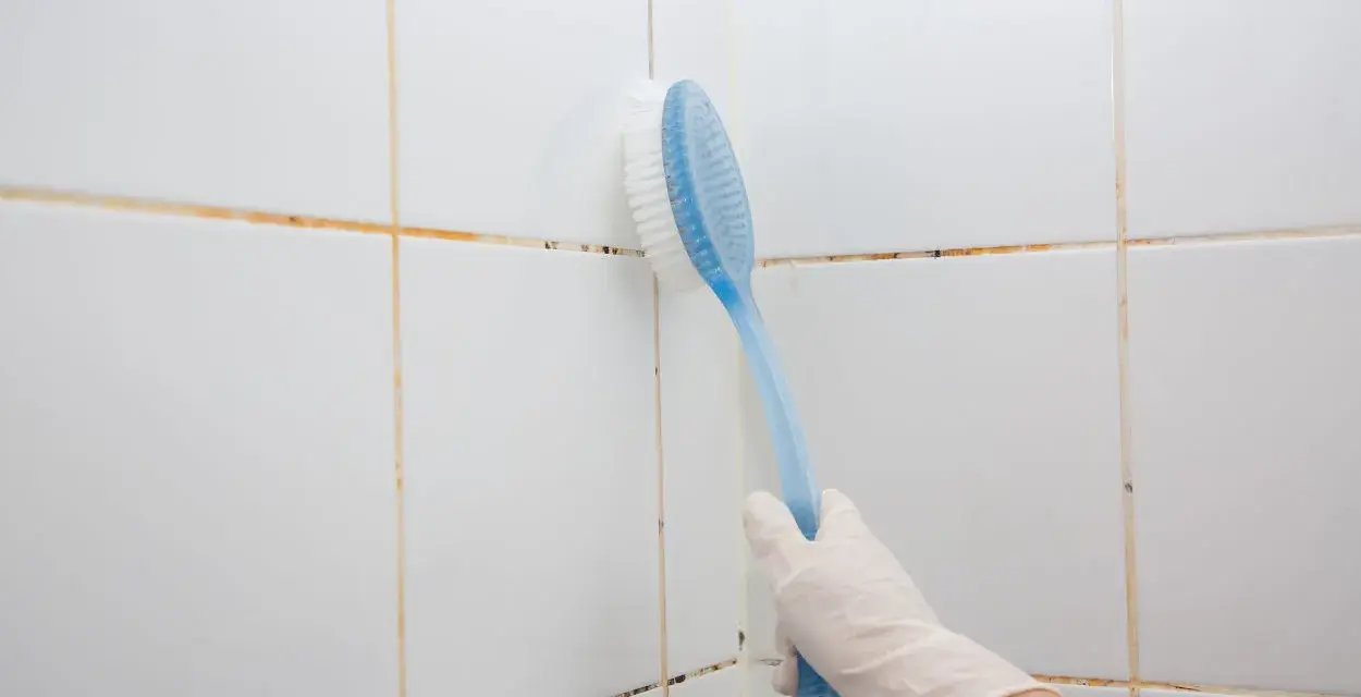 Mano con guanto bianco usa spazzola blu per pulire le fughe delle piastrelle bianche, mostrando come togliere la muffa.