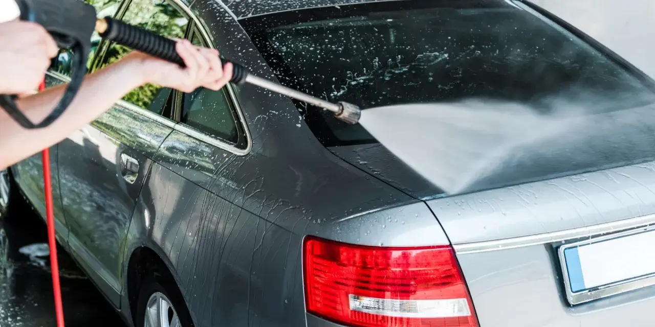 A person demonstrates how to power wash a car, spraying water onto the rear of a grey sedan.