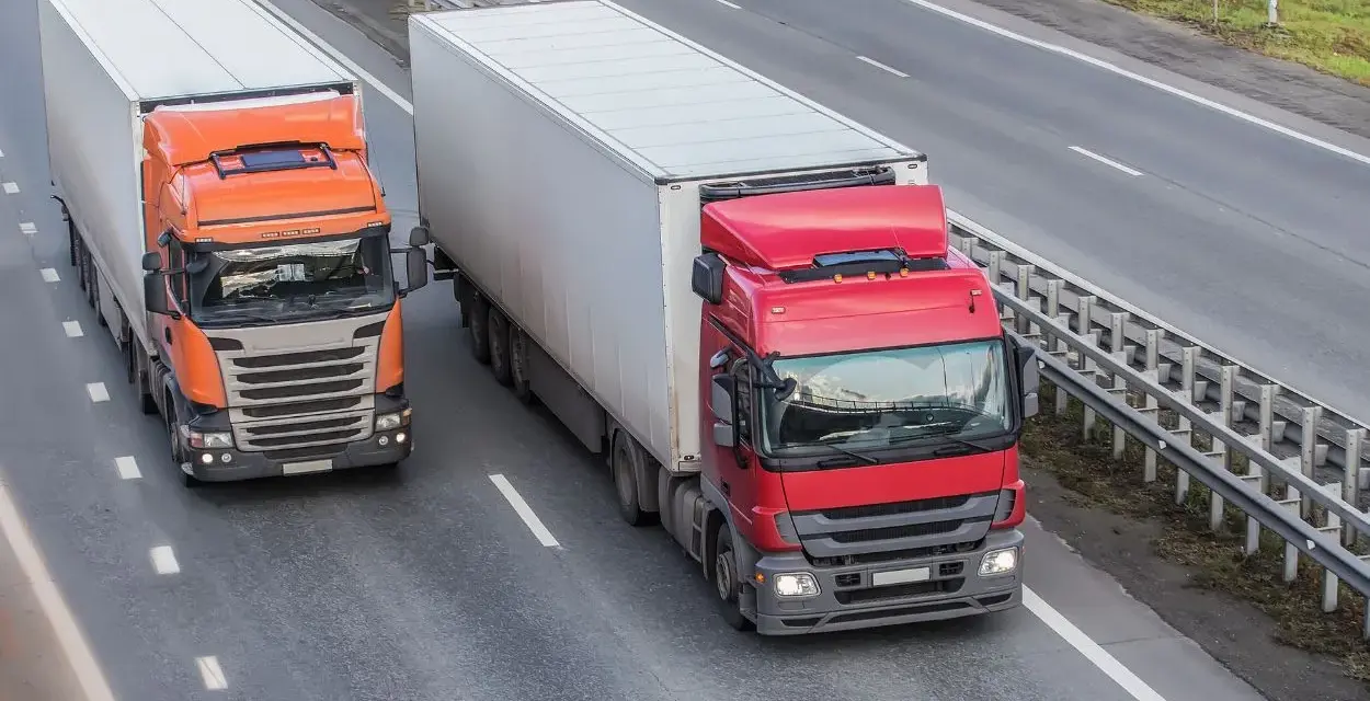 Dwa ciężkie tiry na autostradzie. Czerwony i pomarańczowy, każdy z dużą naczepą. Maksymalny nacisk na oś naczepy to klucz do bezpiecznego transportu.