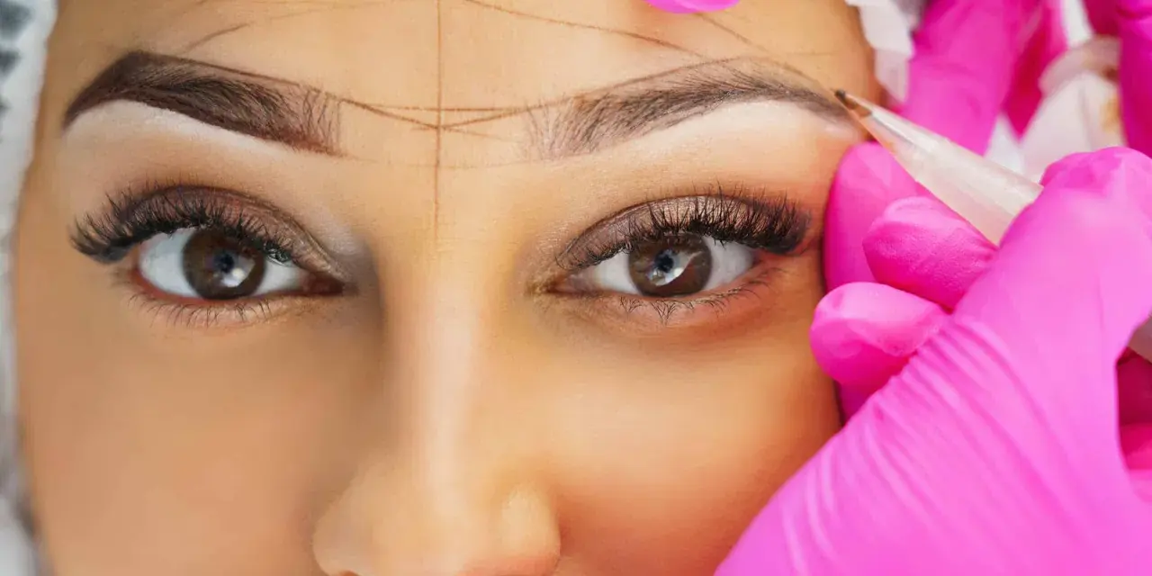 Nahaufnahme einer Frau, deren Augenbrauen mit Linien markiert werden, während eine Person in pinken Handschuhen mit einem Stift arbeitet.