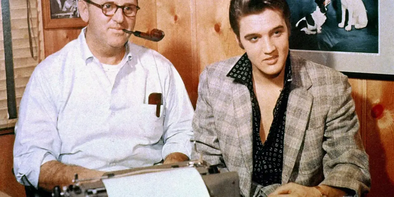 Tom Parker und Elvis Presley sitzen nebeneinander. Parker, mit einer Pfeife im Mund, tippt auf einer Schreibmaschine.
