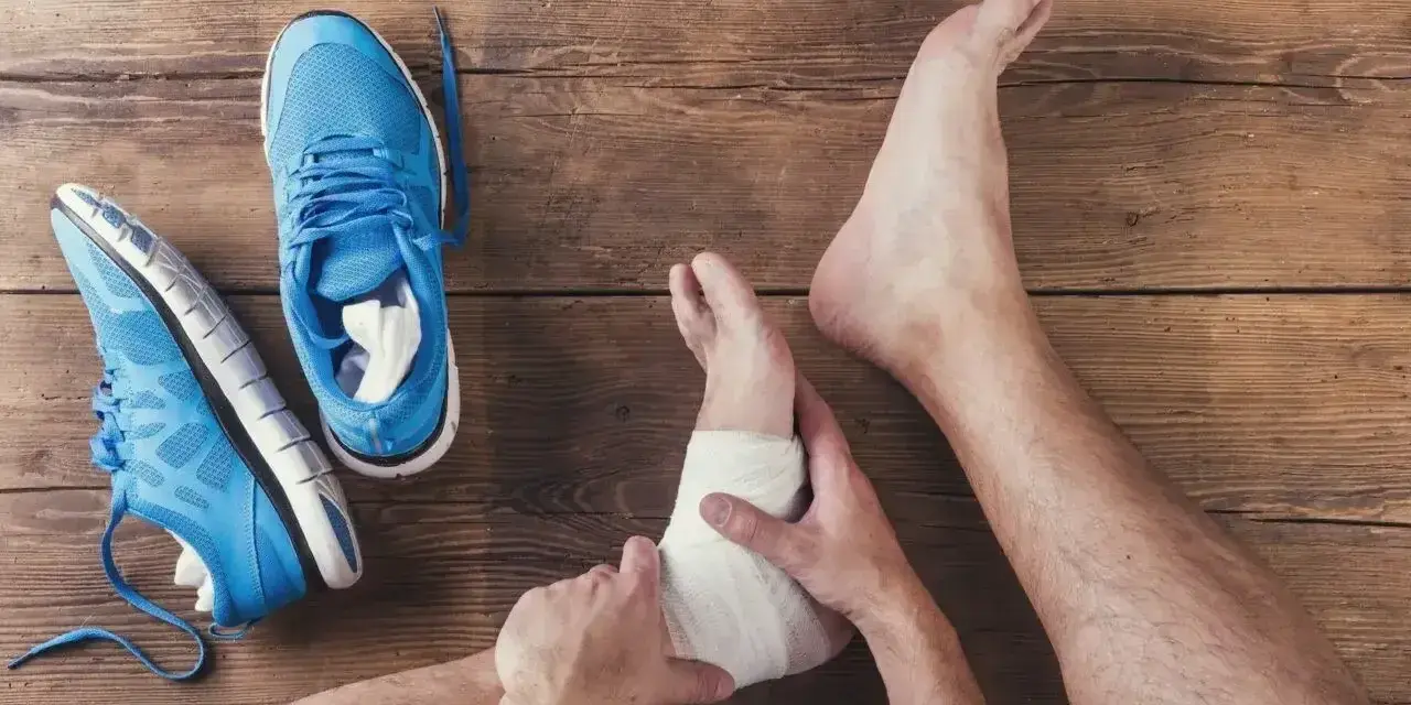 Noga z bandażem na kostce, obok niebieskie buty sportowe. Wciąż widoczna opuchlizna 2 miesiące po skręceniu kostki.