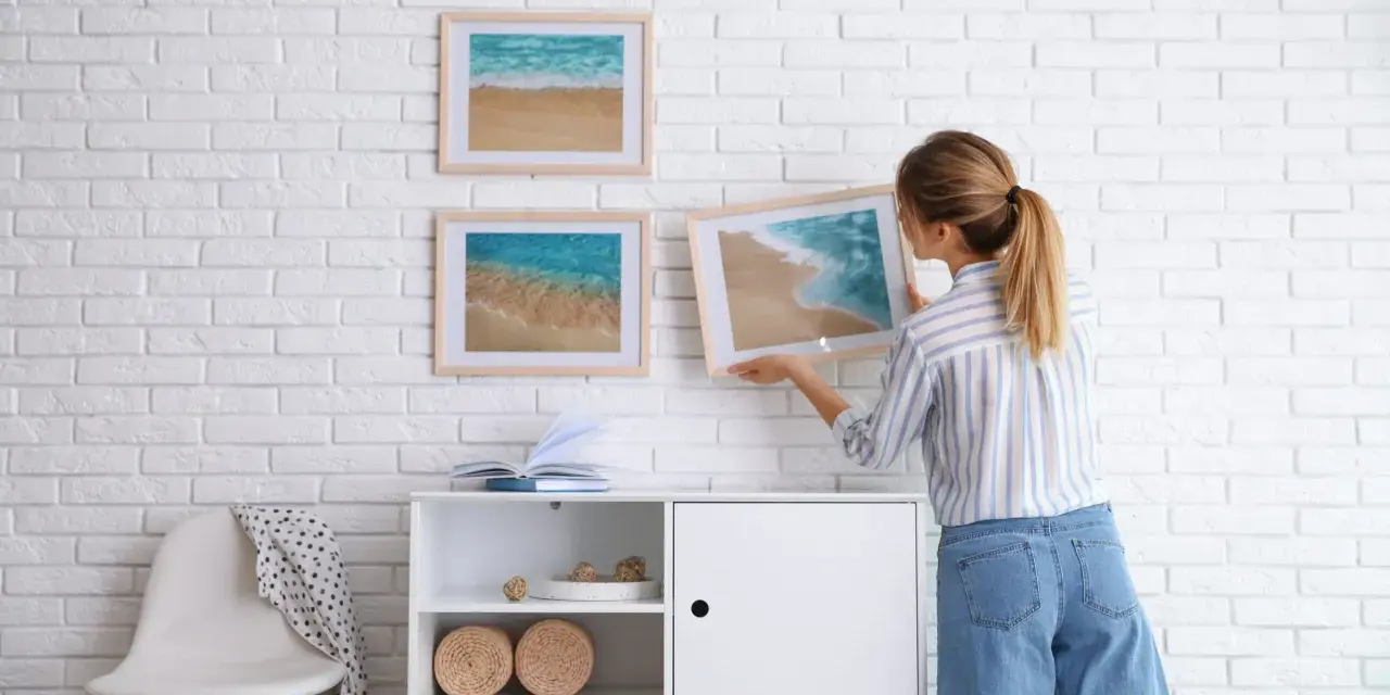 Mujer colocando cuadros de playa en pared de ladrillo blanco. Ideas para como combinar cuadros en la pared.