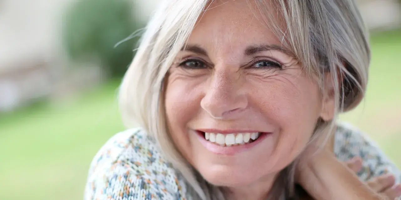 Mujer sonriente con cabello rubio y arrugas normales a los 50 años, disfrutando de un día al aire libre.