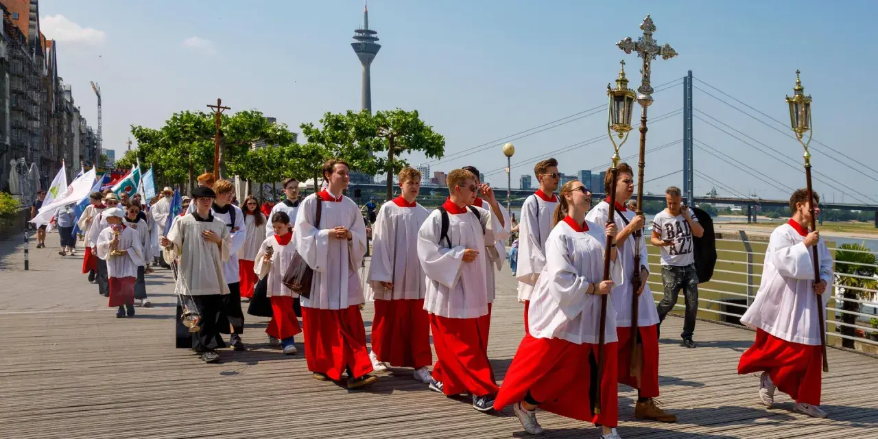 Jugendliche in Soutanen und Alba nehmen an einer Prozession teil, die an christliche Feiertage erinnert. Sie tragen Kreuze und Fahnen.