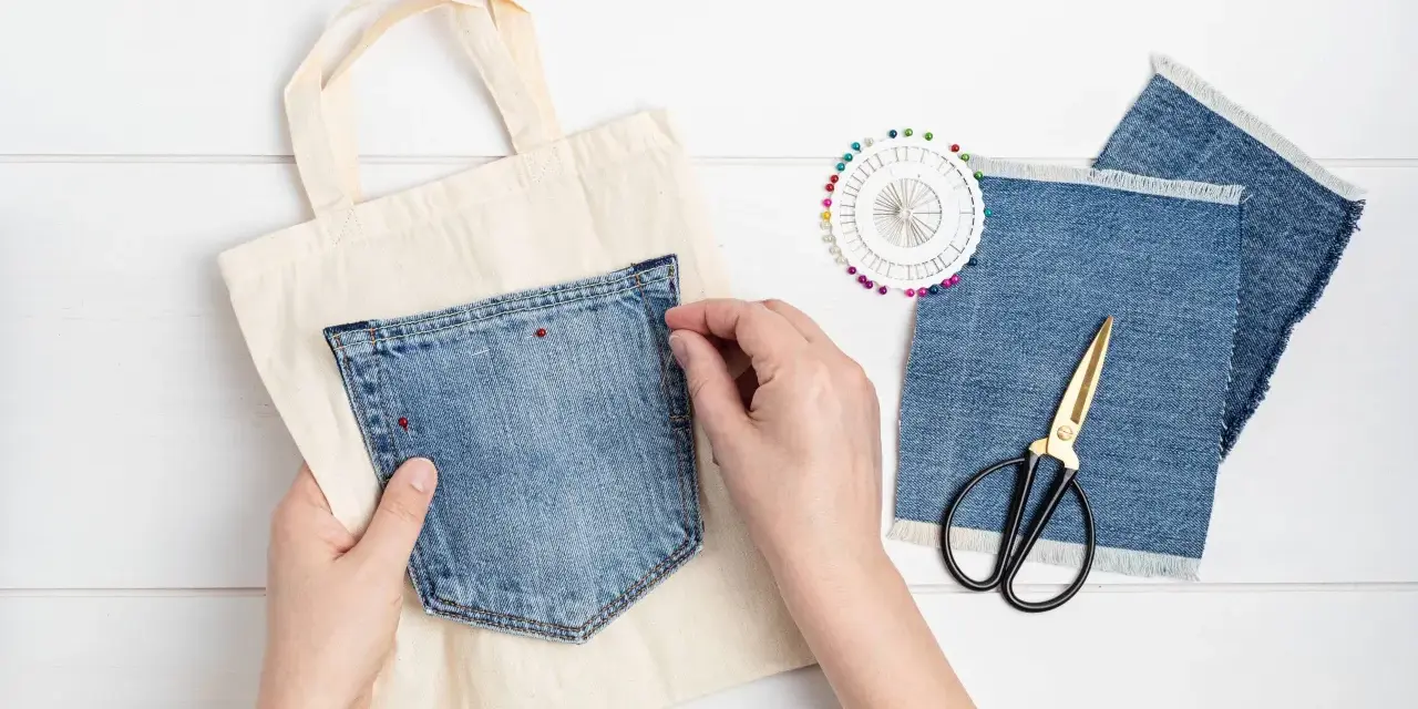 Mains épinglant une poche extérieure en jean sur un sac en toile. Ciseaux et chutes de tissu à proximité.