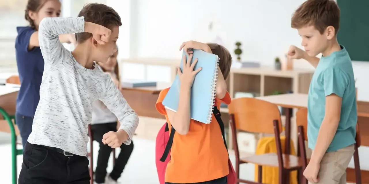 Chłopcy w klasie doświadczają konfliktów między rówieśnikami. Jeden chłopiec grozi pięścią, inny zasłania się książką.