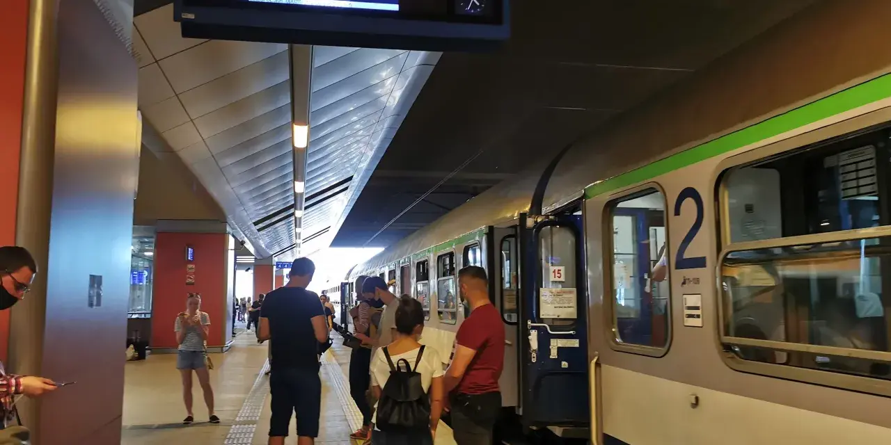 Ludzie na peronie czekają na pociąg, na tablicy informacyjnej widnieje godzina 16:12 i kierunek Gdynia Główna.