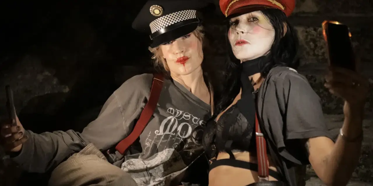 Deux femmes aux maquillages théâtraux portent des casquettes et des bretelles, l'une tenant un téléphone.