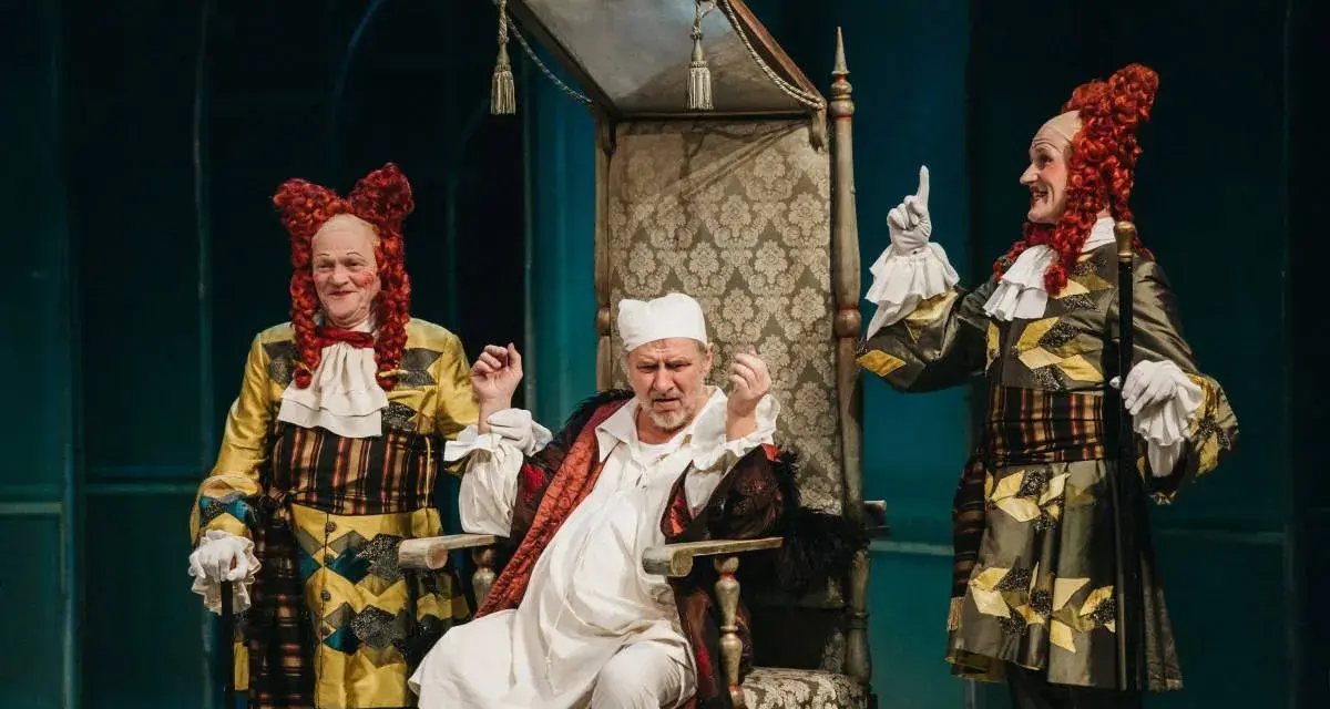 Chory z urojenia siedzi na tronie, otoczony przez dwóch dziwacznie ubranych mężczyzn w perukach.