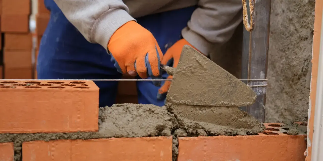 Budowa murowanego prostego grilla z cegły. Ręce w pomarańczowych rękawiczkach nakładają zaprawę kielnią.