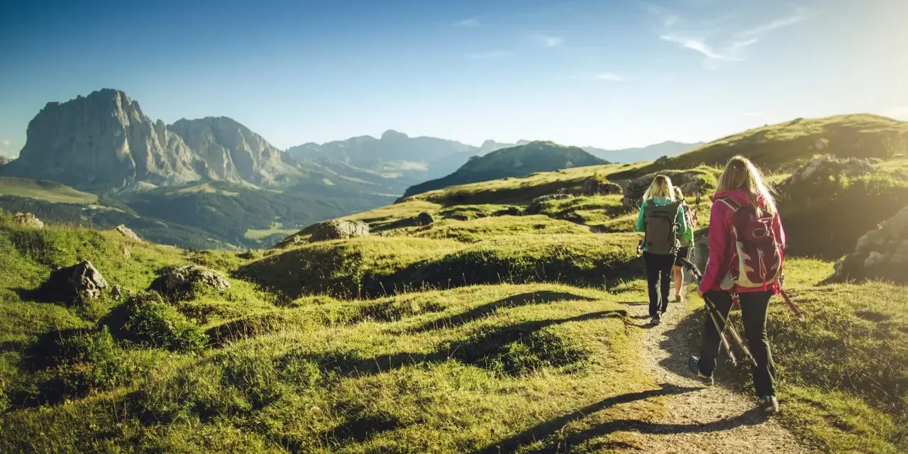 Persone in un percorso di trekking e hiking tra le montagne, con zaini e bastoncini da passeggio, sotto un cielo azzurro.